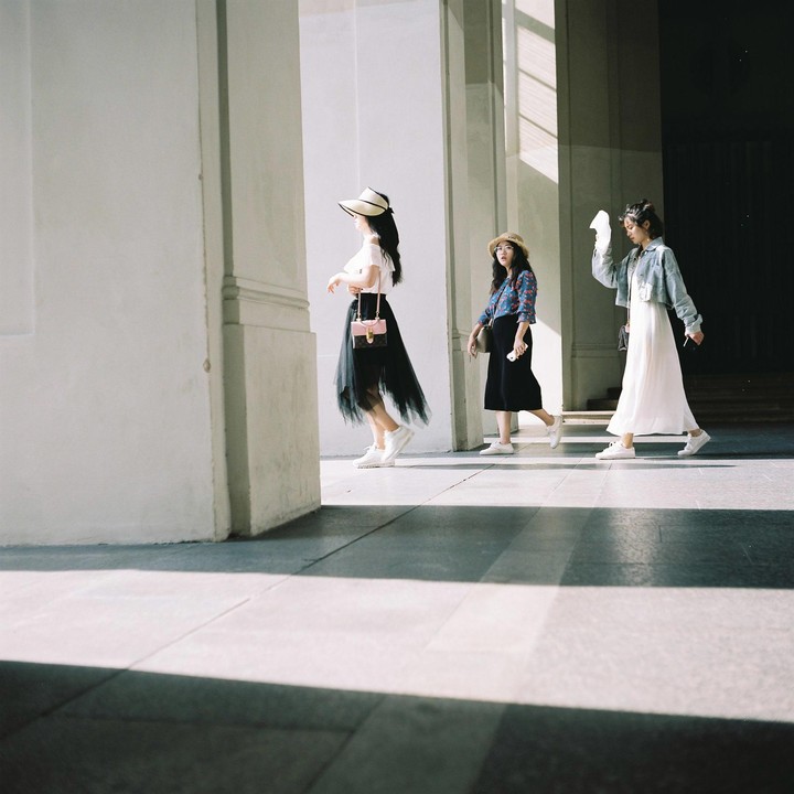 Three women walk along a sunlit corridor with strong shadows.