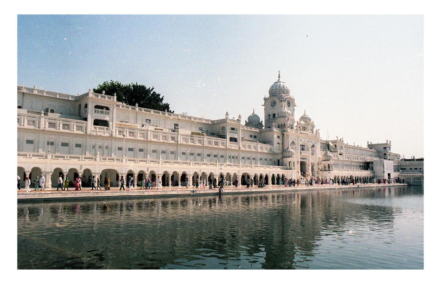 A large white building with a clock tower and domes stands beside a body of water, with many people walking along its arched facade.