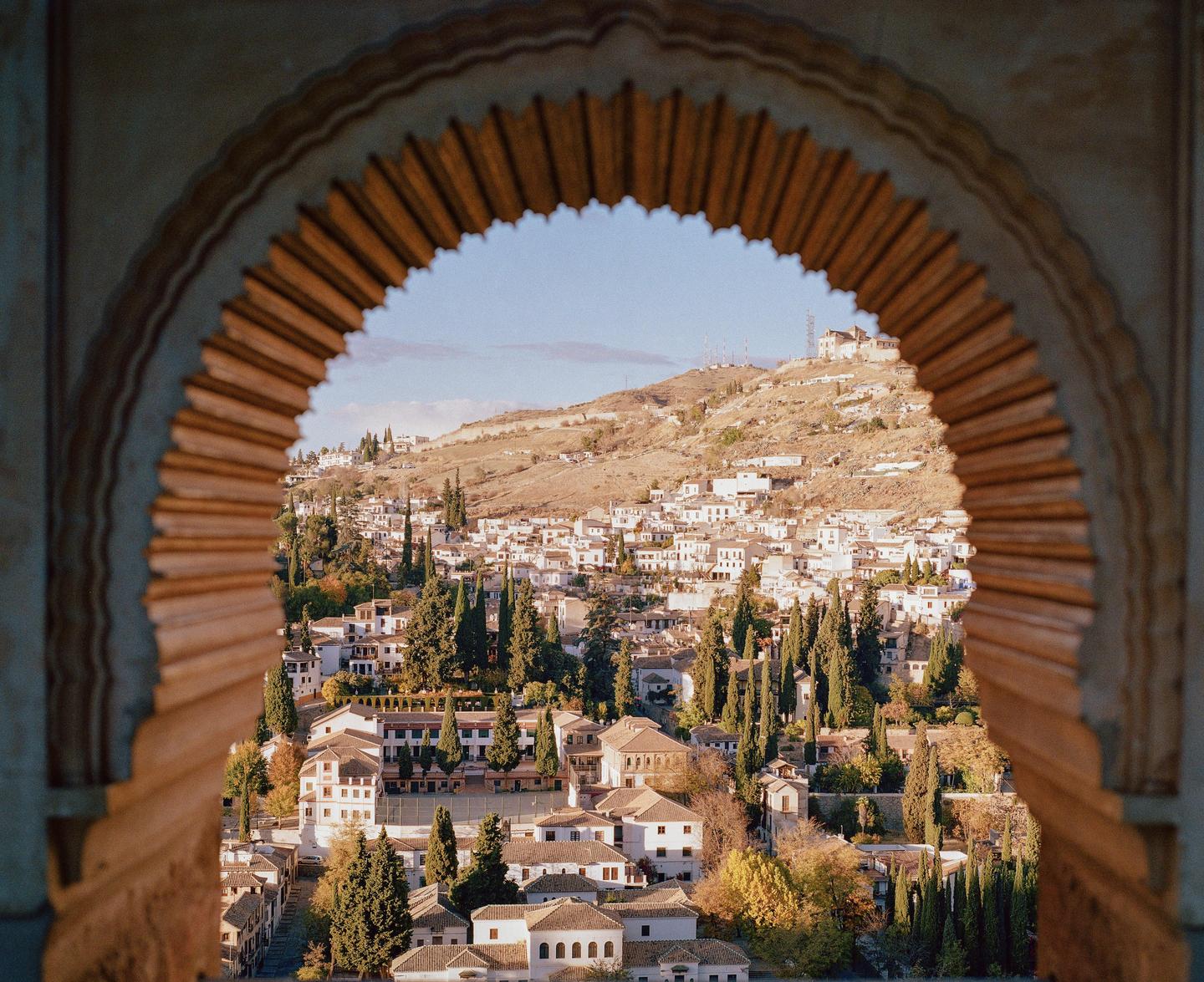 A view of a town with white buildings and cypress trees on a hillside, framed by an ornate archway.