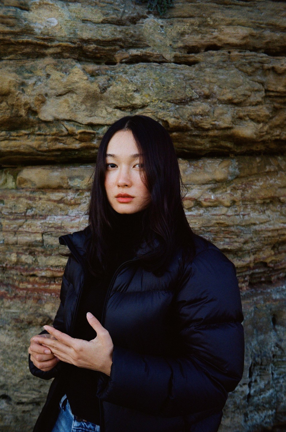 A woman in a black puffer jacket stands in front of a textured stone wall.