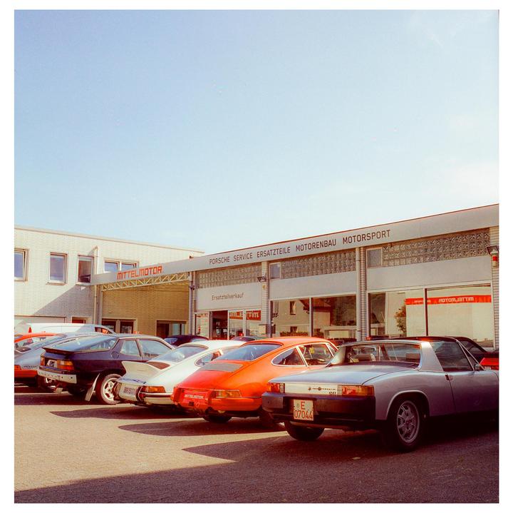 A row of vintage cars is parked in front of a building with a sign that reads "Porsche Service Ersatzteile Motorenbau Motorsport."