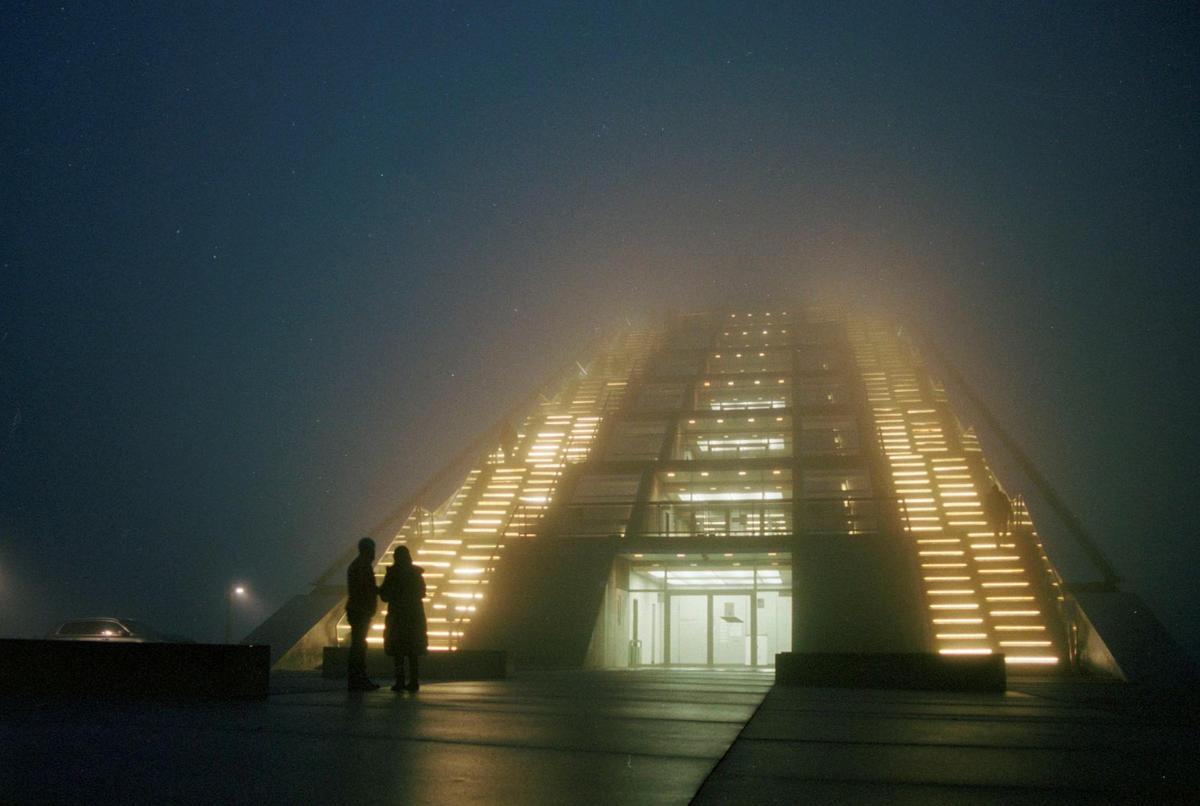 Two people stand in front of a large building with illuminated stairs disappearing into a hazy night sky.