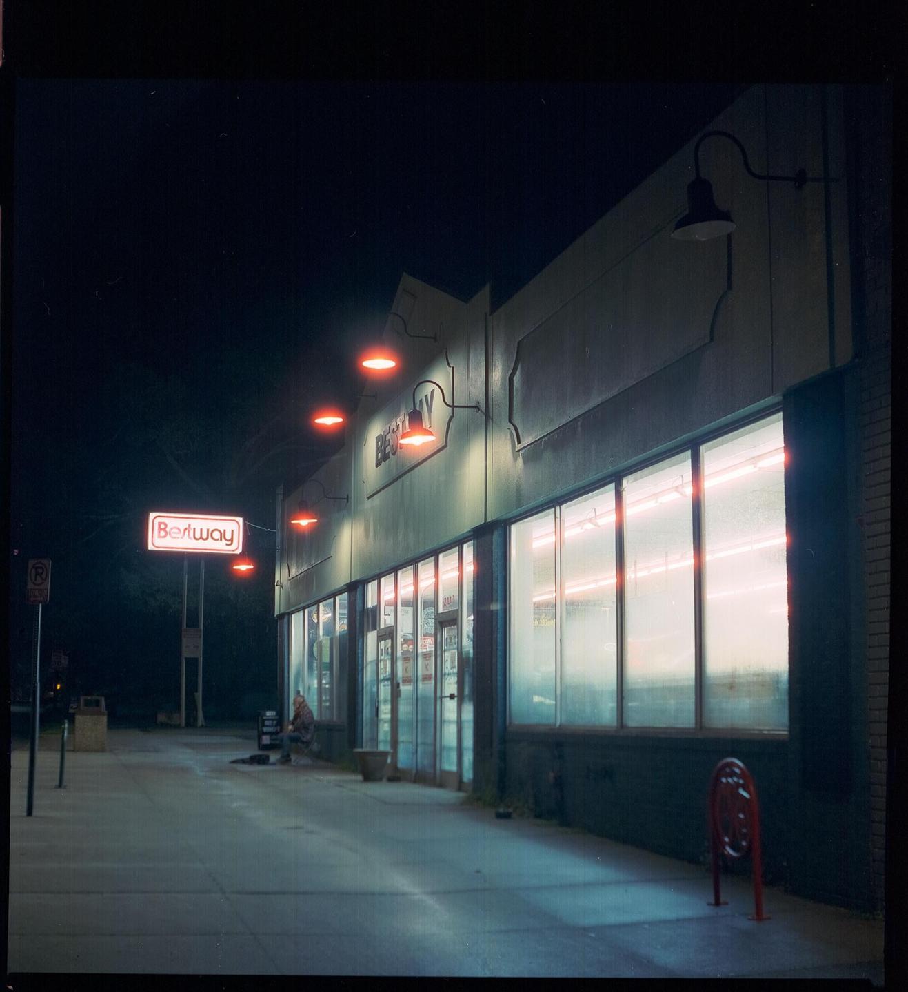 A person sits on the sidewalk outside a store with a neon sign that reads "Berlway" at night.