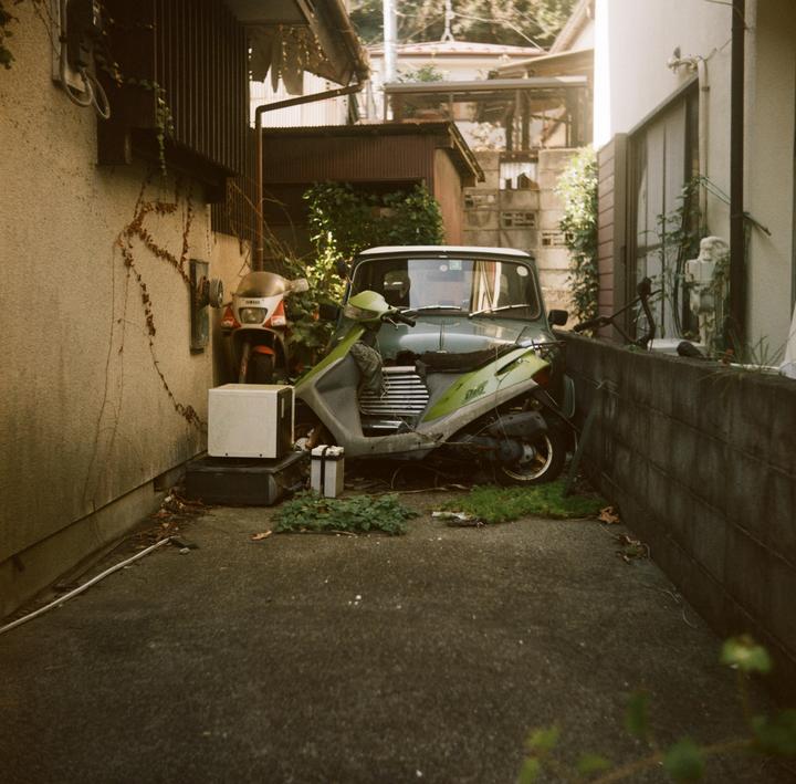 A green scooter and a red motorcycle are parked in a narrow alleyway next to a building.