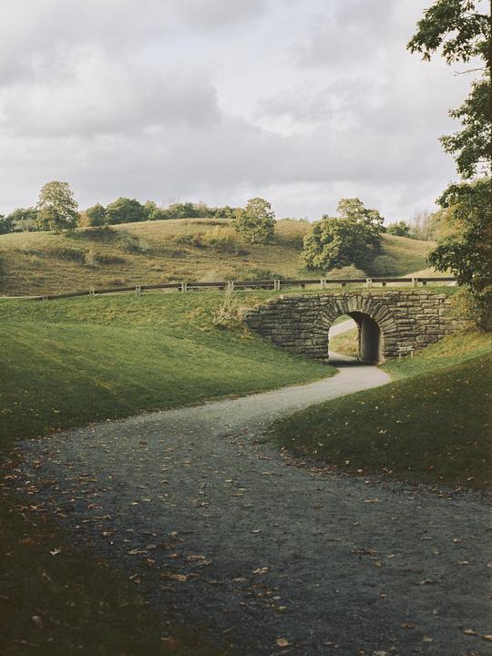 A winding path leads to a stone bridge under a cloudy sky.