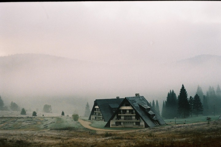 Two A-frame buildings stand in a field with a forest and mountains in the background, all shrouded in fog.