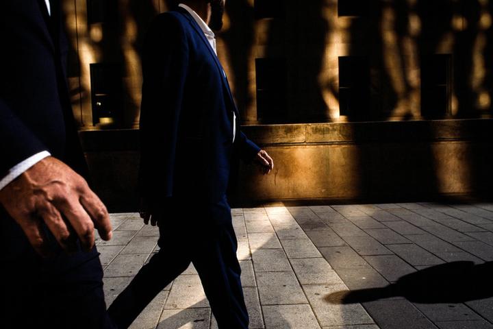 Two men in suits walk on a sidewalk with strong shadows and light patterns on a building wall.