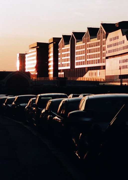 A row of dark cars is parked in front of modern buildings illuminated by the setting sun.