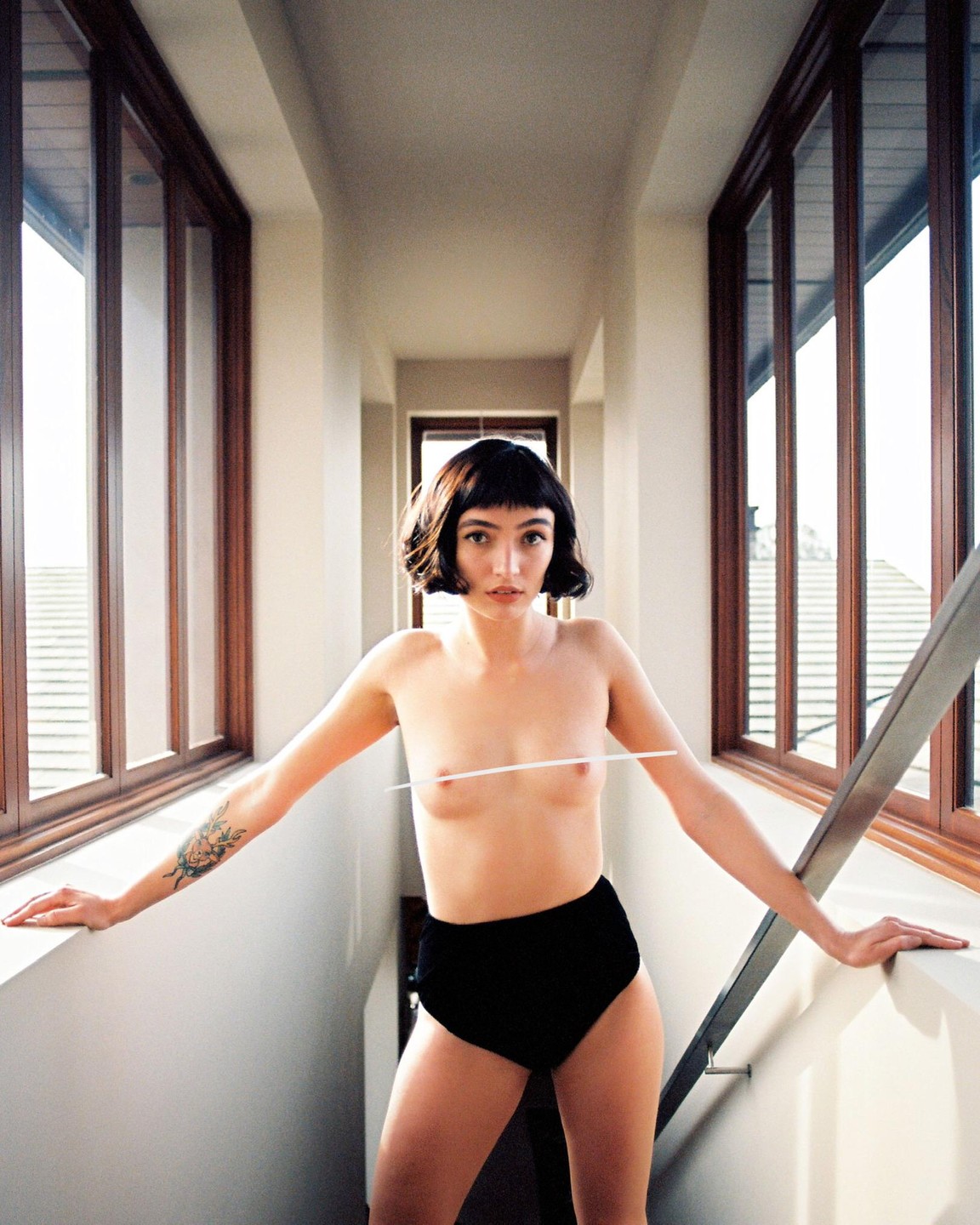 A topless woman with short dark hair and black underwear stands in a sunlit hallway with large windows, holding onto a railing.