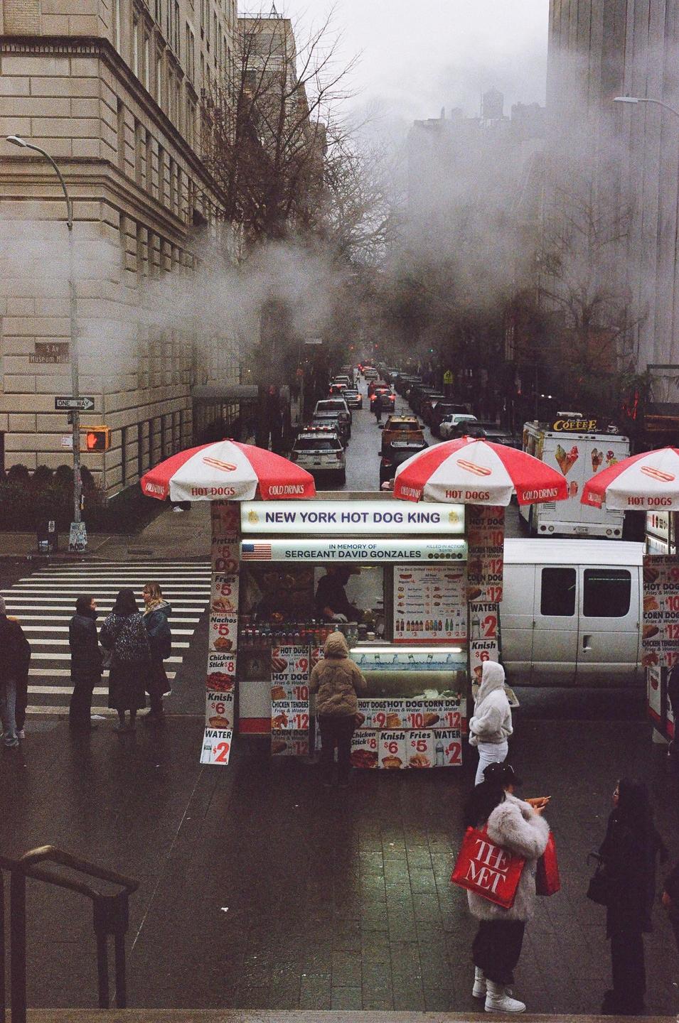 A street vendor sells hot dogs from a cart with red and white umbrellas on a city street with cars and pedestrians.