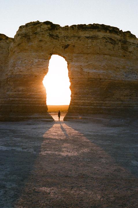 A person walks through a natural rock archway with the setting sun visible in the distance.