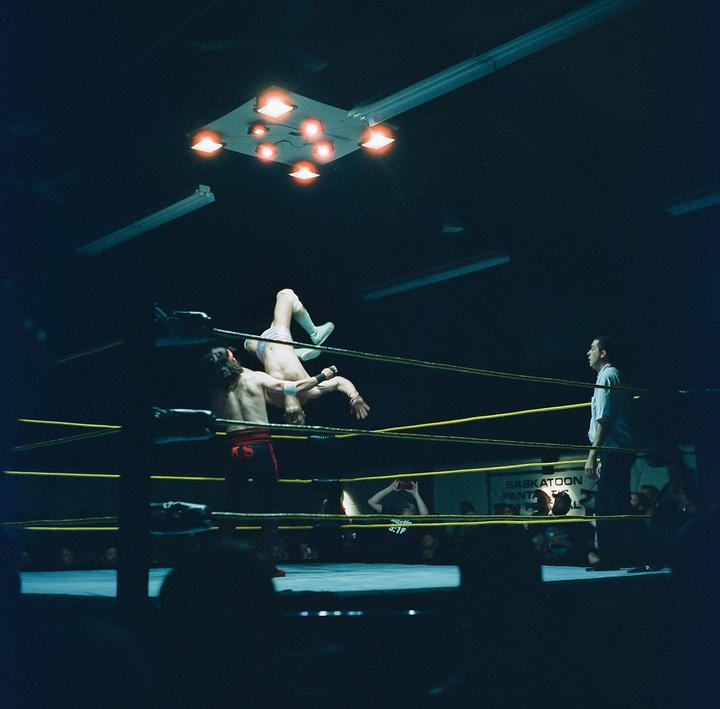 Two men wrestle in a ring while a third man watches and an audience looks on from the dark.
