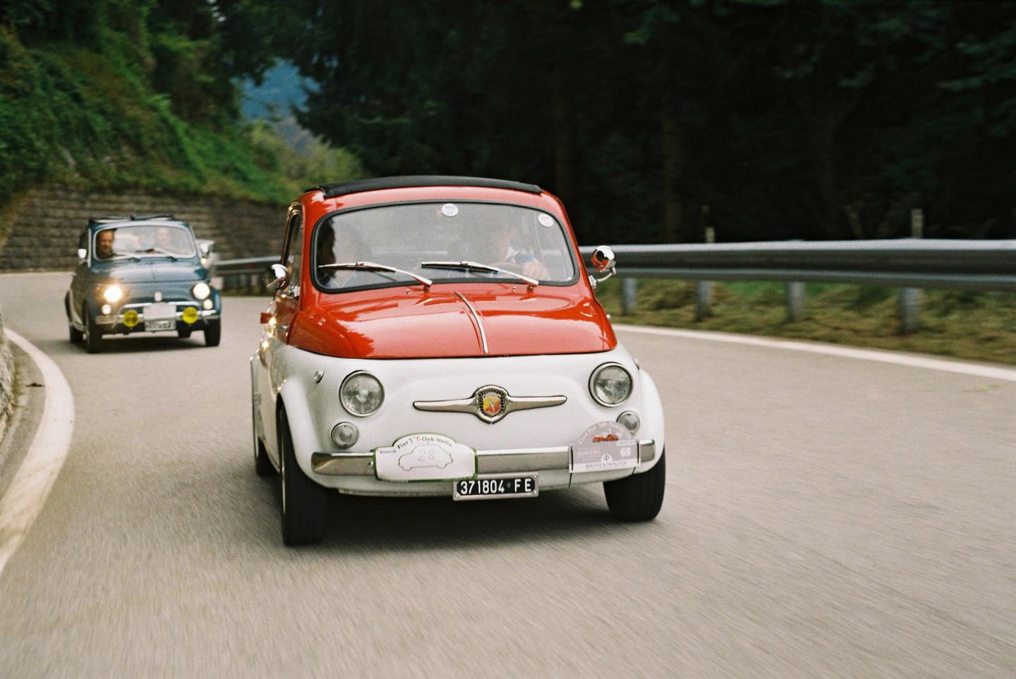 Two Fiat 500 cars drive on a winding road through the alps.