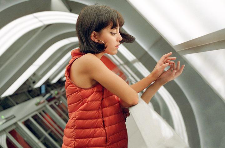 A woman in an orange vest leans on a railing with her hands together, looking down, with a background of curved architectural elements.