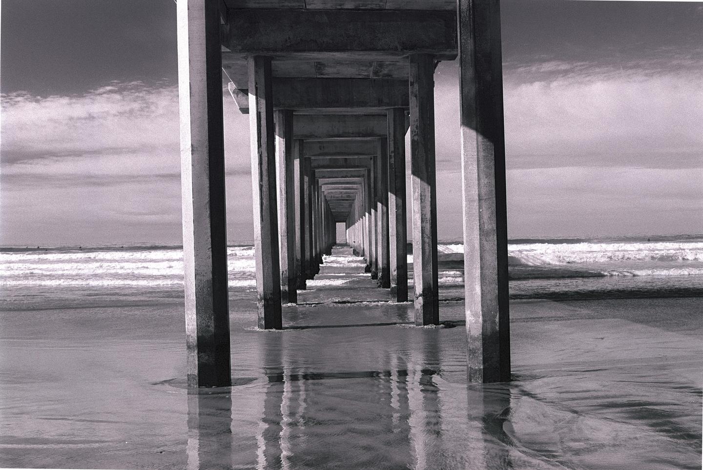 The underside of a pier with many concrete pillars extends into the distance over a sandy beach with waves.