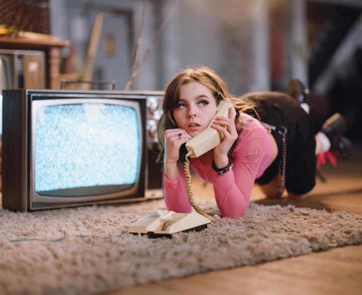 A young woman in a pink top lies on a carpet, holding a landline phone to her ear, with a static-filled television screen beside her.