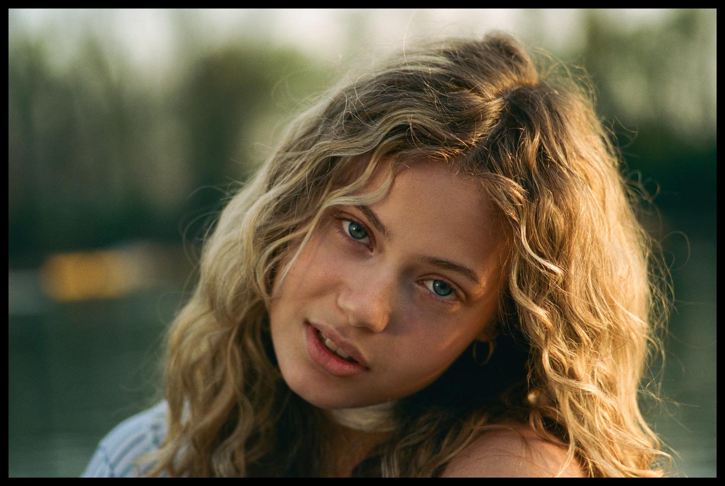 A young woman with curly blonde hair and blue eyes looks directly at the viewer.