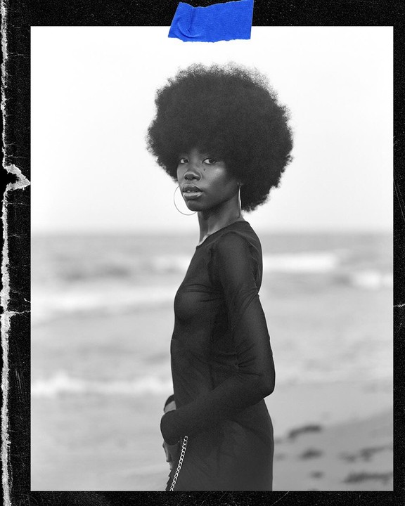 A woman with an afro and hoop earring stands on a beach.