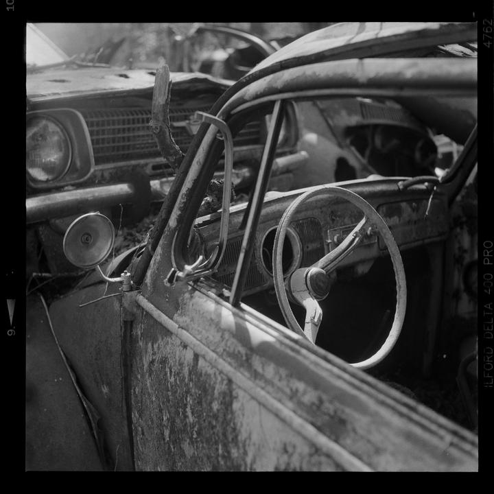 A black and white photograph shows the interior and exterior of a rusty, abandoned car with another car visible in the background.