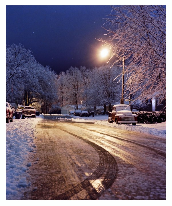A snow-covered street at night is illuminated by a street light, with cars parked along the side and tire tracks visible in the snow.