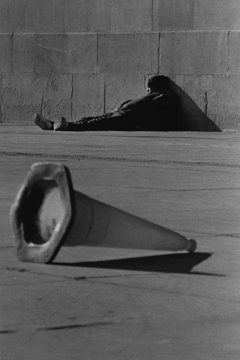 A man rests against a wall while a traffic cone lies on the ground in the foreground.