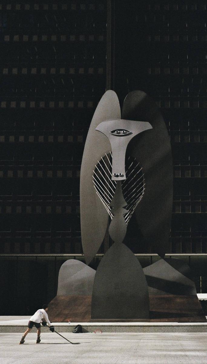 A person plays street hockey in front of a large Picasso sculpture.