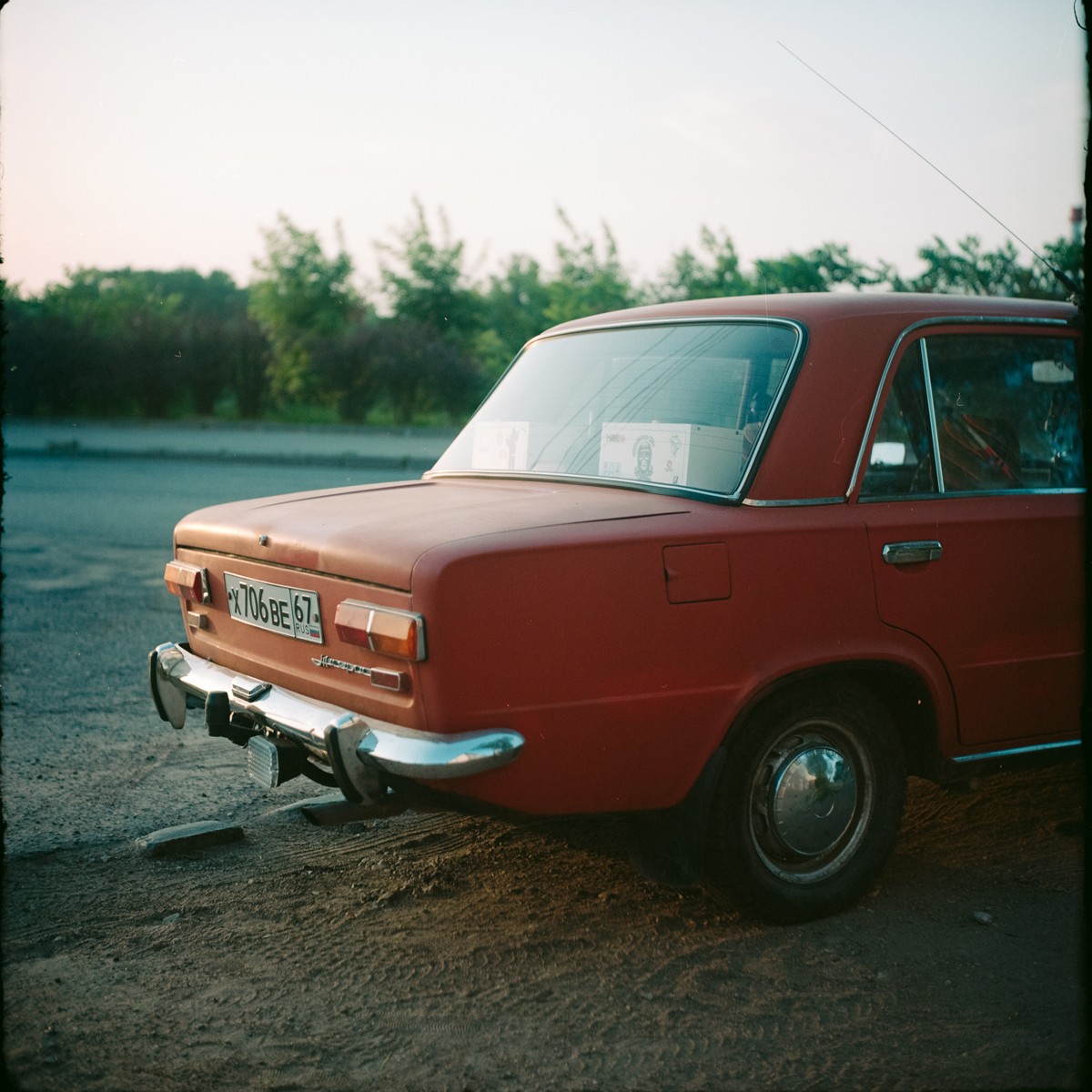 A red car is parked on a dirt and asphalt surface with trees in the background.