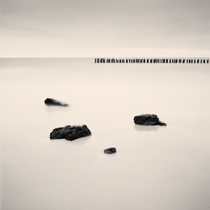 Four dark rocks are visible in the foreground of a calm body of water, with a line of wooden posts in the distance under a hazy sky.