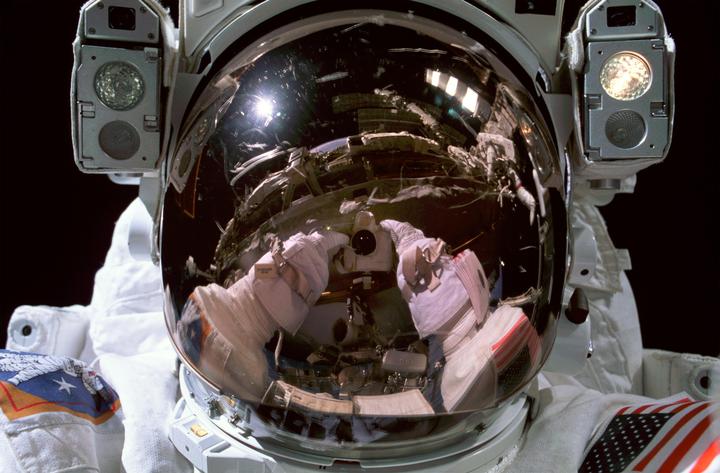 An astronaut in a space suit takes a self-portrait with a camera, reflected in the helmet's visor.