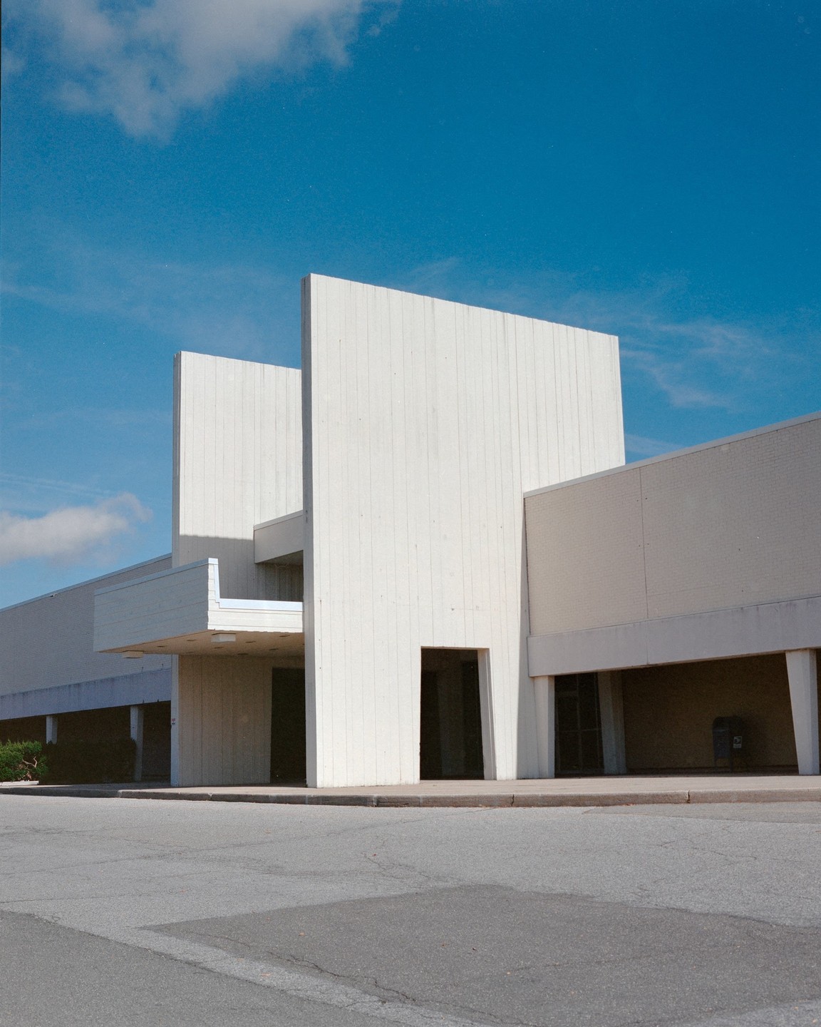 A large, minimalist building with tall, flat white walls and dark entrances stands under a bright blue sky with a few clouds.