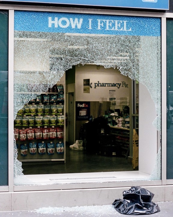 A storefront window is shattered, with a large hole revealing the interior of a pharmacy.
