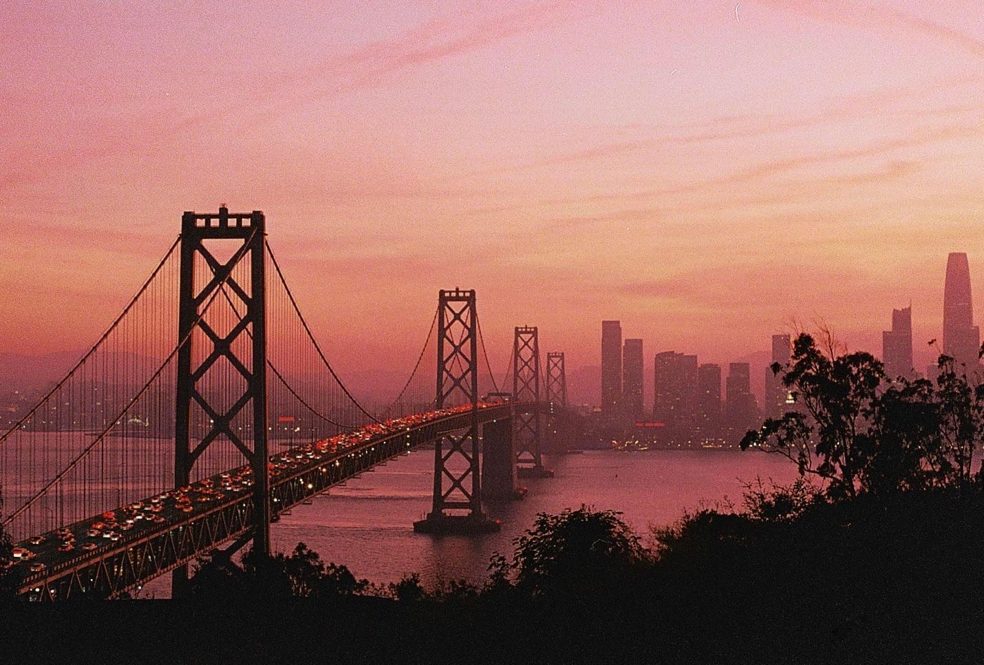 A suspension bridge with heavy traffic crosses a body of water towards a city skyline under a pink and orange sunset.