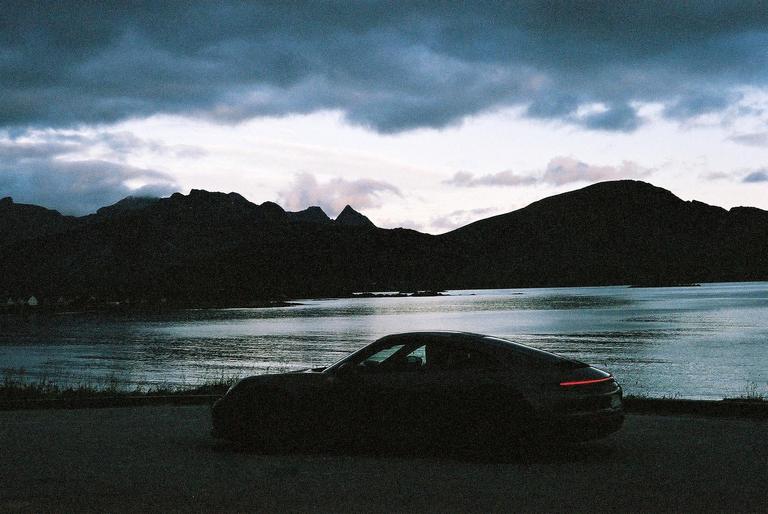 A dark car is parked on a road overlooking a body of water with mountains in the background under a cloudy sky.