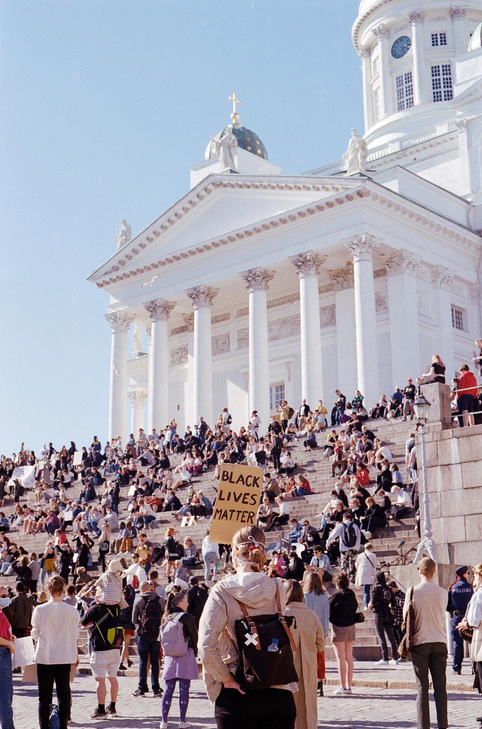 A crowd of people gathers on the steps of a large white neoclassical building, with one person holding a "Black Lives Matter" sign.