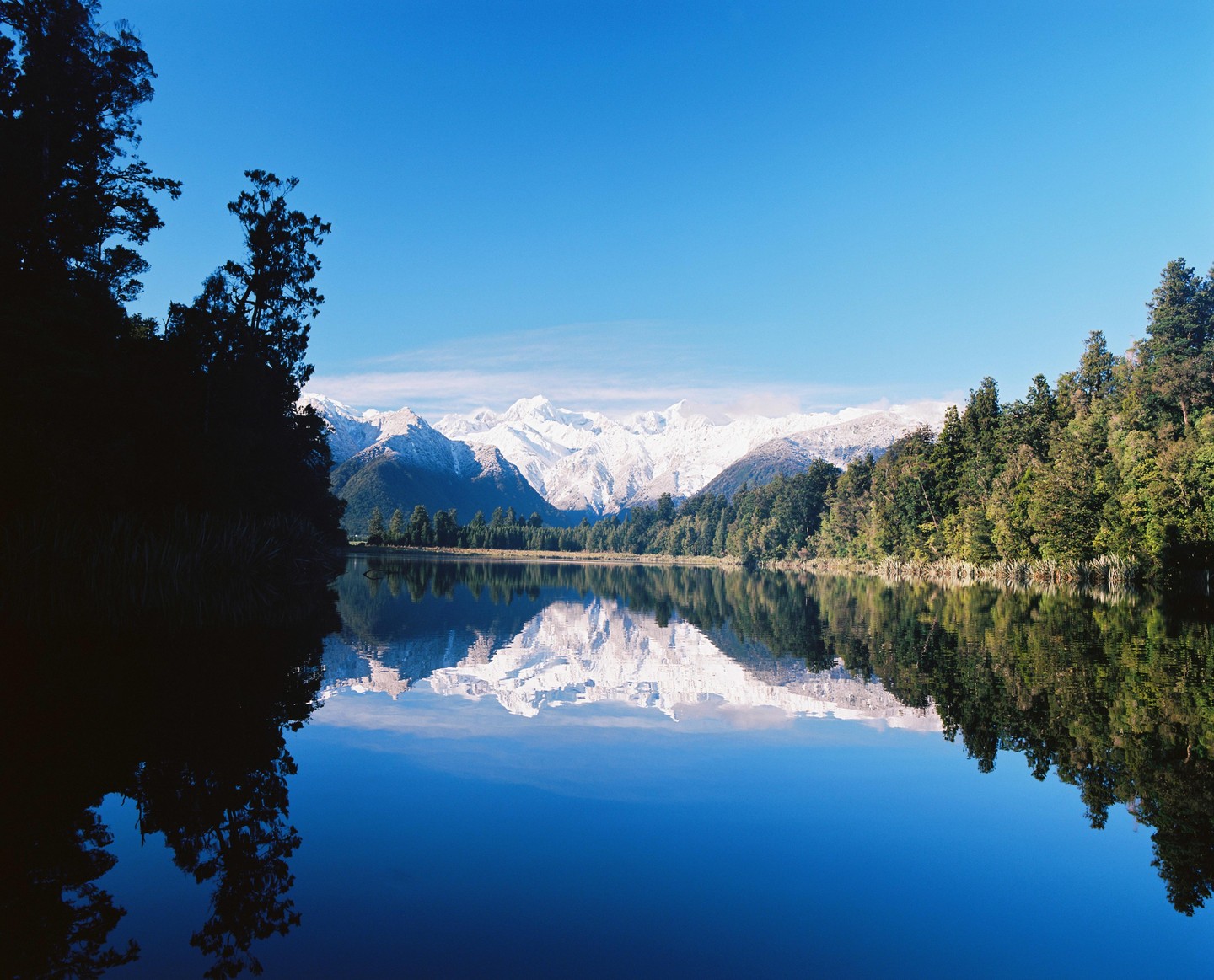 A calm lake reflects snow-capped mountains and a forest under a clear blue sky.