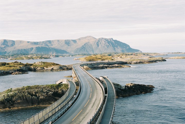 A winding road bridge stretches across a body of water, connecting rocky islands with mountains in the background.