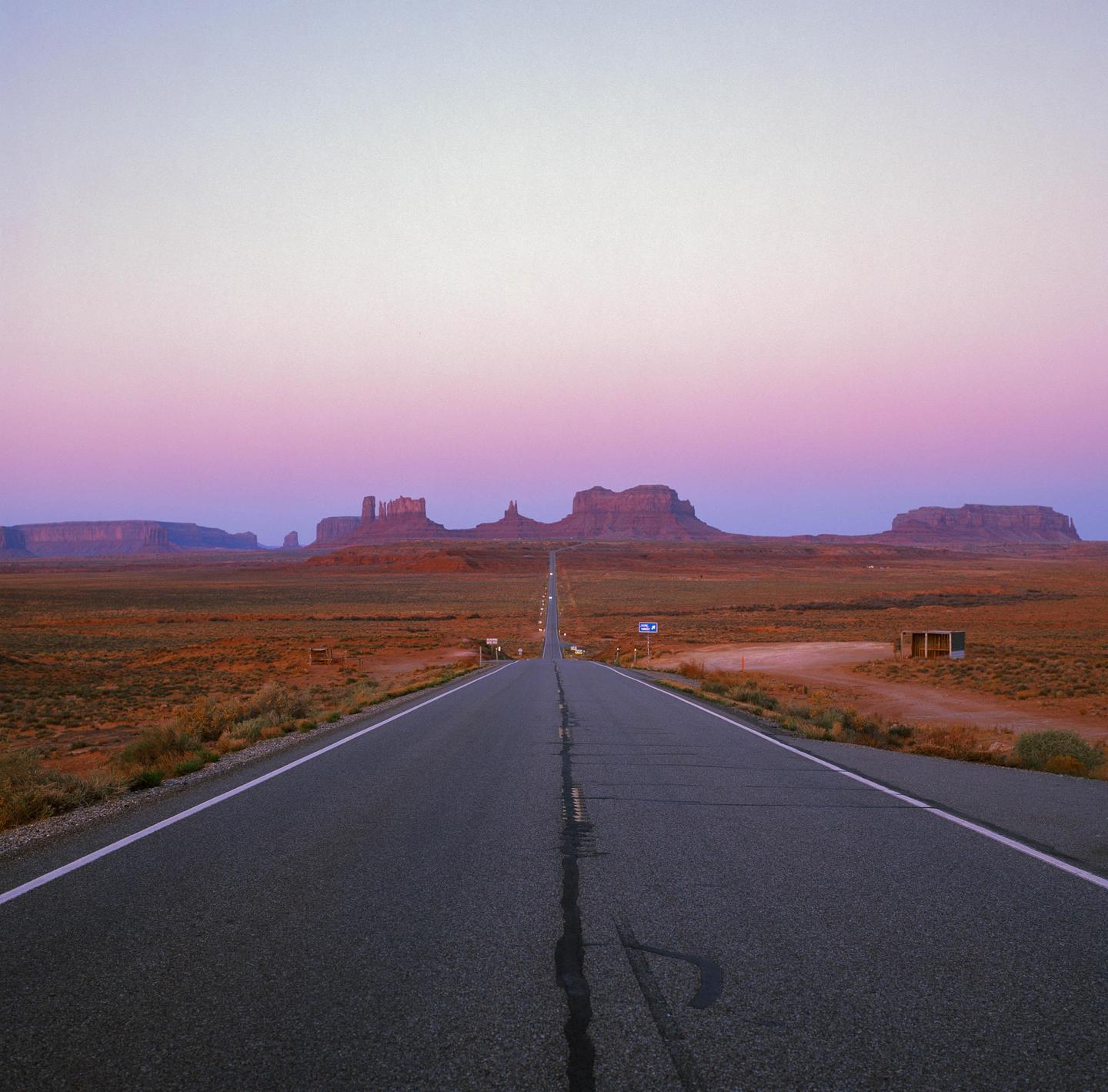 A long, straight road leads towards distant mesas and buttes under a colorful sky at dusk.