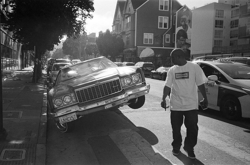 A man walks past a lowrider car parked on a city street with one wheel lifted.