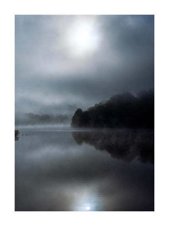 A misty lake reflects the bright sun and dark trees under a cloudy sky.