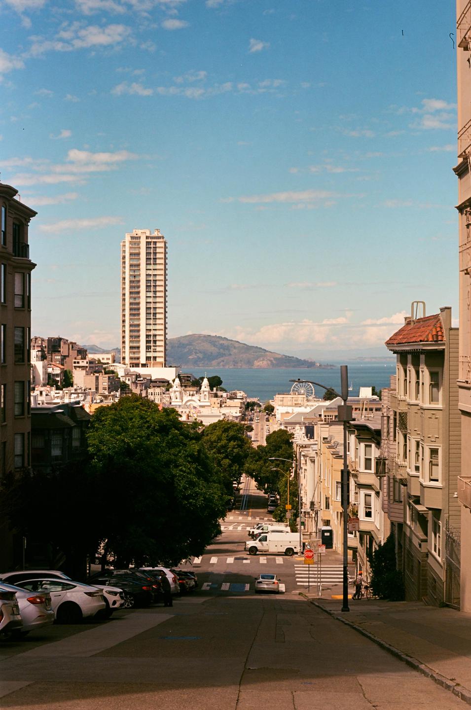 A tall building stands prominently in a city with a bay and mountains in the distance.