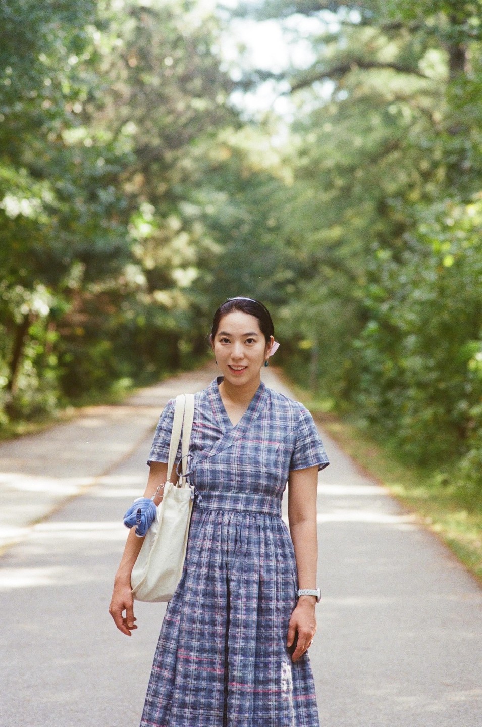A woman in a plaid dress stands on a tree-lined road, holding a tote bag.