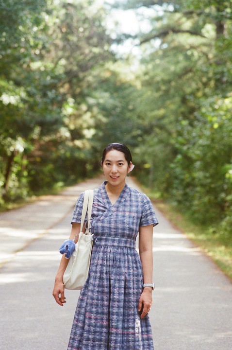 A woman in a plaid dress stands on a tree-lined road, holding a tote bag.