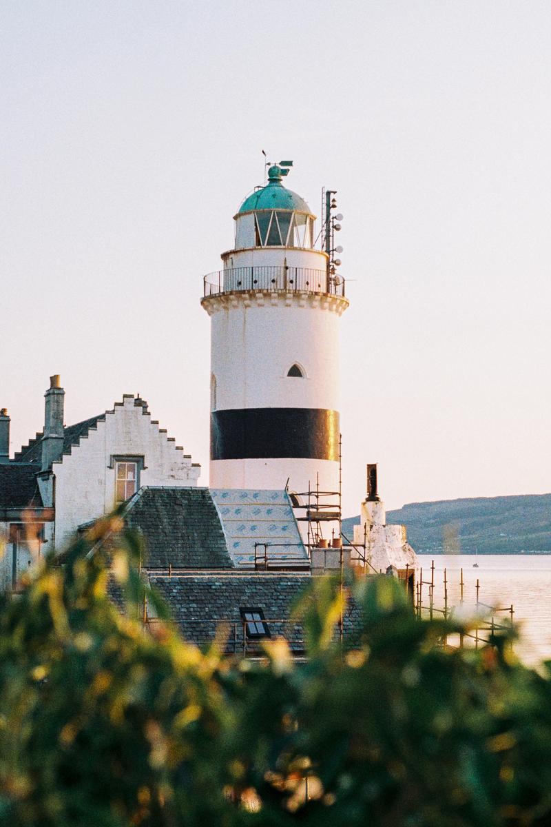 A white lighthouse with a black stripe and a green top stands next to a building with scaffolding, overlooking water and distant hills.