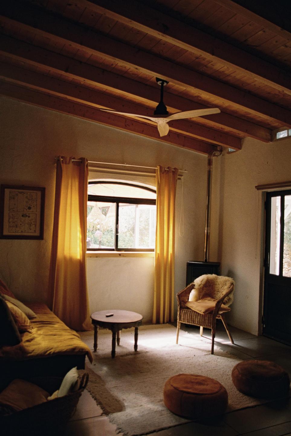 Sunlight streams through a window with yellow curtains into a rustic room with a wooden ceiling, a sofa, a wicker chair, and a small table.