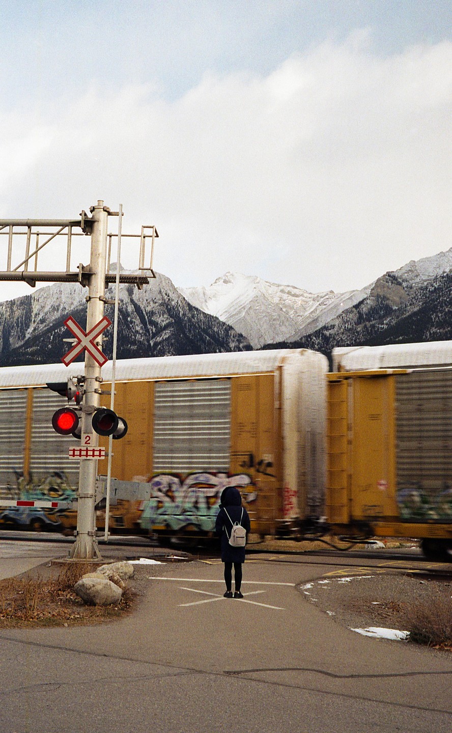 A person stands at a railroad crossing while a train passes, with snow-capped mountains in the background.