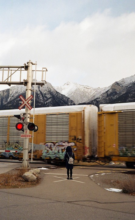 A person stands at a railroad crossing while a train passes, with snow-capped mountains in the background.