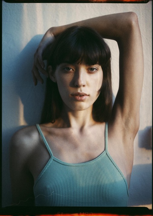 A woman with dark hair and bangs wears a light green tank top and poses with one arm raised above her head.