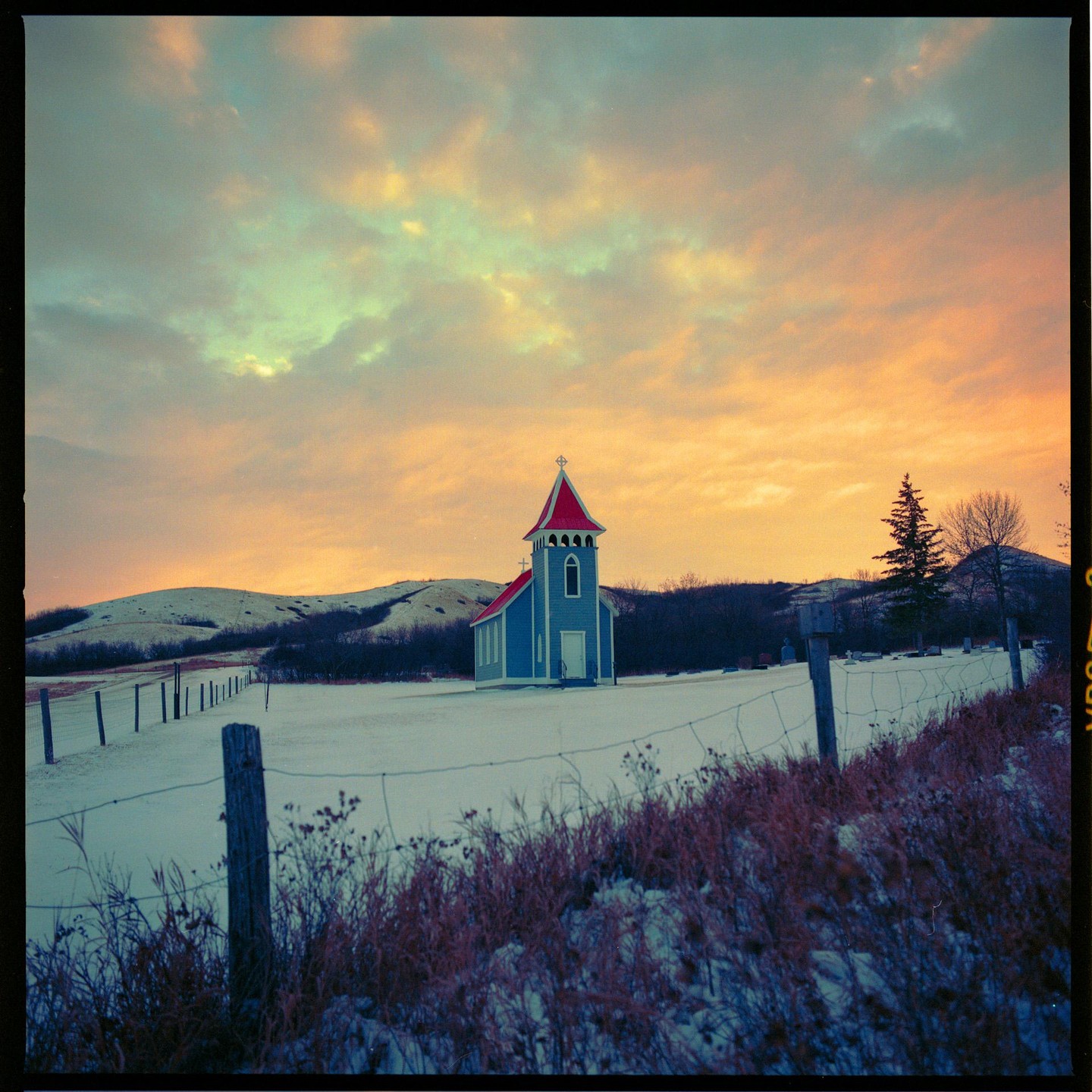 A small blue church with a red roof stands in a snow-covered field under a colorful sky.