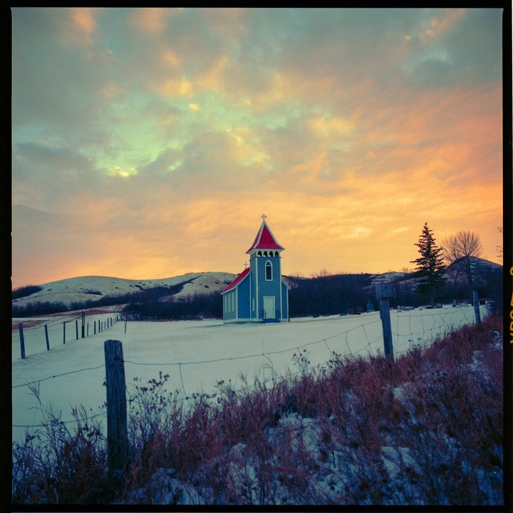A small blue church with a red roof stands in a snow-covered field under a colorful sky.