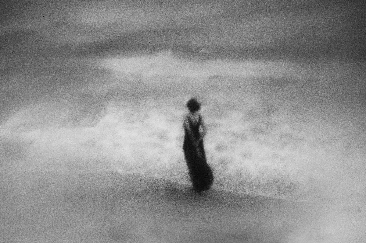 A woman stands in the water at the edge of a body of water, with waves breaking around her.